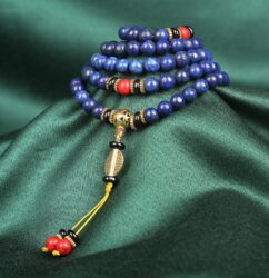 Multi-layered lapis lazuli beaded bracelet with gold, black, and red accent beads, featuring a decorative gold pendant and tassel with red and black beads on a green fabric background.