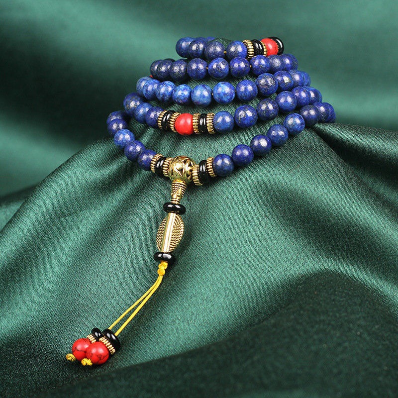 Multi-layered lapis lazuli beaded bracelet with gold, black, and red accent beads, featuring a decorative gold pendant and tassel with red and black beads on a green fabric background.